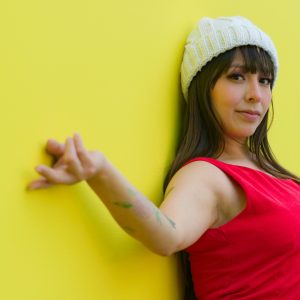 Stylish woman wearing red dress and beanie posing against a vibrant yellow wall.